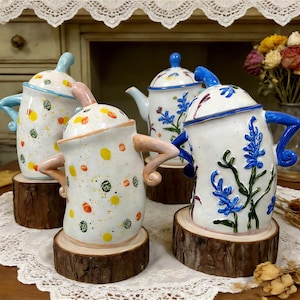 Könnte beinhalten: Vier Keramik-Teekannen mit einzigartigen Designs. Zwei Teekannen haben bunte Tupfen, während die anderen beiden blaue Blumenmuster aufweisen. Jede Teekanne steht auf einer Holzscheibe und präsentiert eine skurrile und künstlerische Ästhetik. Die Teekannen haben Griffe und Deckel.