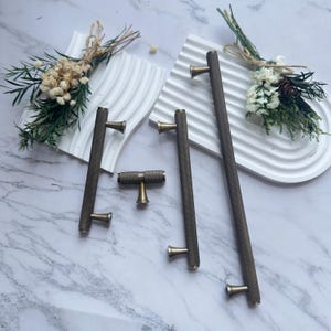 Pode incluir: Uma coleção de ferragens para armários em tons de bronze, incluindo puxadores longos e um botão em forma de T. A ferragem apresenta uma superfície texturizada com padrão de diamante e detalhes em cor de latão. Os itens estão dispostos em uma superfície de mármore.