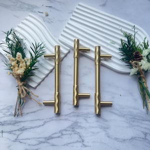 May include: Three gold-colored cabinet pulls with a geometric design. Each pull has a T-shaped handle and a faceted body. The pulls are arranged on a marble surface with white decorative trays and floral accents.