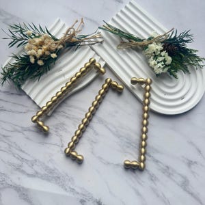 May include: Three gold-colored cabinet pulls with a beaded design are displayed on a marble surface. Two white, arched trays with dried flowers and greenery are also present. The pulls are in a modern style.