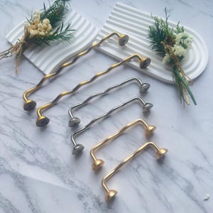 Puede incluir: Un conjunto de tiradores de armario metálicos ondulados en tonos dorados y plateados. Los tiradores tienen un diseño texturizado único y están dispuestos sobre una superficie blanca con detalles florales decorativos. Estos tiradores decorativos son ideales para armarios.