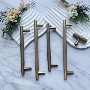 May include: A collection of bronze-colored cabinet pulls and knobs. The pulls have a textured, diamond-patterned grip and a T-shaped design. The hardware is displayed on a white, wavy surface with floral accents, showcasing a modern aesthetic.