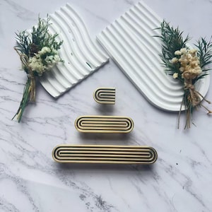May include: A collection of gold-toned cabinet hardware with a black accent, including a small knob, a medium-sized pull, and a long pull. The hardware is arranged on a marble surface with white, arched decorative trays and small bouquets.