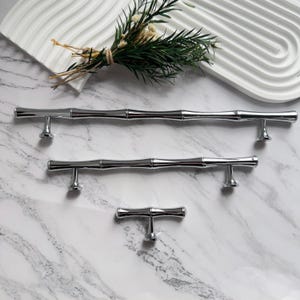 May include: Three polished chrome cabinet pulls in varying sizes. The pulls have a bamboo-like design, with a series of segmented sections. The hardware is displayed on a marble surface, with a decorative white tray and greenery in the background.