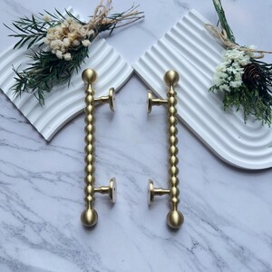 May include: Two brass door handles with a beaded design. Each handle has a round ball at the top and bottom, with a series of smaller spheres in between. The handles are mounted on a marble surface.