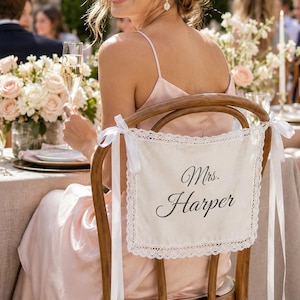 Puede incluir: Un letrero rectangular de tela color hueso con el texto en cursiva negra Mrs. Harper. El letrero está bordeado por un delicado encaje blanco y se sujeta al respaldo de una silla de madera con cintas de satén blanco atadas en las esquinas.