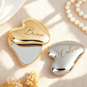 May include: Two heart-shaped compact mirrors with metallic finishes. One mirror is gold-toned with the name Brian engraved in script, and the other is silver-toned with the name Calvin engraved in script. Both items feature a smooth, reflective surface.