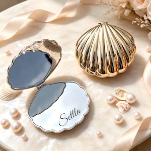 May include: A gold-coloured, shell-shaped compact mirror with a hinged design. One unit is closed, while the other is open, revealing two reflective surfaces. The bottom interior mirror features the name 'Setlla' in a black, cursive script font.
