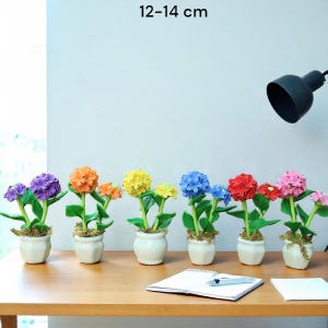 以下が含まれることがあります： 小さな白い鉢に入った、カラフルな造花の紫陽花のコレクション。花は紫、オレンジ、黄色、青、赤、ピンクの色合いです。各植物の高さは約12〜14cmです。植物は木製の表面に配置されています。