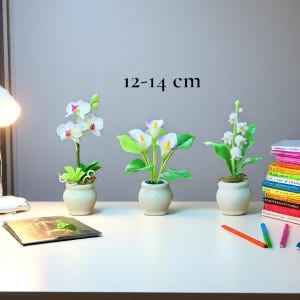May include: Three miniature artificial flower arrangements in small cream-coloured pots. The arrangements include an orchid, calla lilies, and lily of the valley. The height of the arrangements is 12-14 cm.