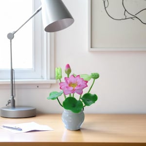 May include: A small, white vase holding a pink lotus flower arrangement with green leaves and buds. The artificial flowers are displayed on a wooden desk, next to a silver desk lamp and a piece of paper.