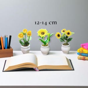 Pode incluir: Três plantas artificiais de girassol em vasos de cerâmica brancos. Os girassóis têm pétalas amarelas brilhantes e folhas verdes. Os vasos estão dispostos sobre uma superfície branca com um livro aberto e um porta-lápis de madeira. O texto "12-14 cm" está acima das plantas.