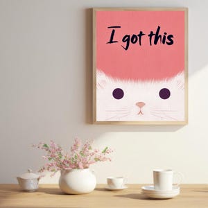 May include: A framed print with a pink background and the text "I got this" in black. A white cat face with black eyes is peeking out from behind the text.