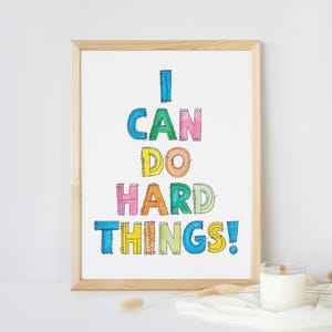 以下が含まれることがあります： カラフルな輪郭のある文字で「I CAN DO HARD THINGS!」と書かれた額装されたプリント。文字はピンク、ブルー、グリーン、イエロー、オレンジのさまざまな色合いで塗りつぶされています。