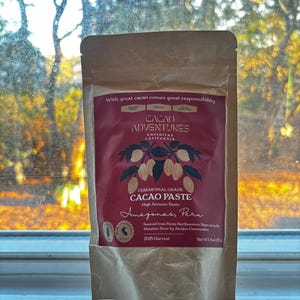 以下が含まれることがあります： Cacao Adventuresの儀式用カカオペーストが入った茶色の紙袋。袋には、白い文字とカカオポッドのイラストが入ったバーガンディ色のラベルが付いています。テキストには「Amazonas, Peru」と「Net WT 8 oz (227g)」と書かれています。