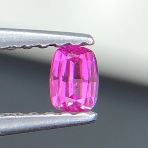 May include: A bright pink, cushion-cut gemstone is held by silver tweezers. The gemstone is rectangular with rounded corners and multiple facets, reflecting light. The background is a soft gradient of grey and white.