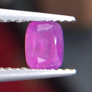 Puede incluir: Una gema de color rosa púrpura, tallada en forma de cojín, es sostenida por unas pinzas plateadas. La superficie facetada de la gema refleja la luz, mostrando su claridad y color. El fondo está borroso.