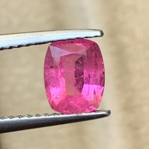 May include: A vibrant, cushion-cut, pink gemstone is held by a pair of tweezers. The gem has a rectangular shape with rounded corners and multiple facets that reflect light. The color is a bright, saturated pink.