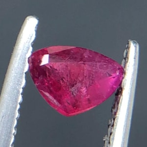 Puede incluir: Un rubí triangular facetado, sostenido por unas pinzas plateadas. El rubí es de un color rojo-rosado intenso, con inclusiones internas visibles. Las pinzas están enfocadas, resaltando la forma y el color de la gema.