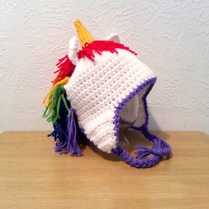 May include: White crocheted unicorn hat with a rainbow mane and purple tassel. The hat has a pointed ear and a horn.