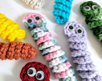 May include: A collection of crocheted worm toys with googly eyes. The worms are in various colors, including yellow, orange, red, purple, blue, green, and pink.