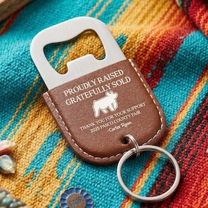 Pode incluir: Um abridor de garrafa de couro marrom com um abridor de metal prateado. O abridor tem o texto "PROUDLY RAISED GRATEFULLY SOLD" e "THANK YOU FOR YOUR SUPPORT 2025 PASCO COUNTY FAIR - Carter Ryan". Um porta-chaves prateado está preso.