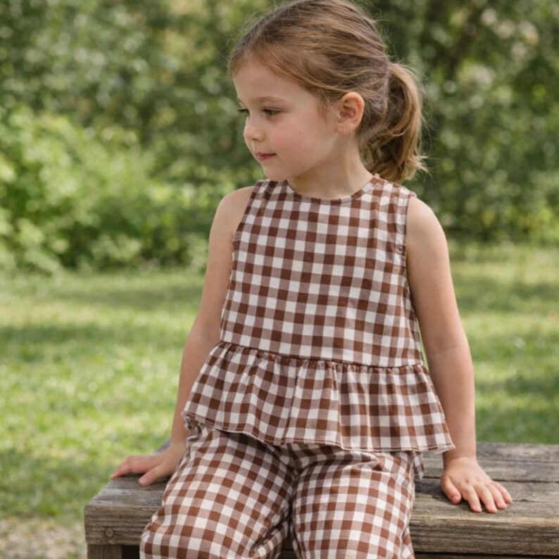 May include: A young child wearing a brown and white checkered sleeveless top and matching pants. The outfit has a ruffled hem on the top. The child is sitting on a wooden bench outdoors with a blurred green background.