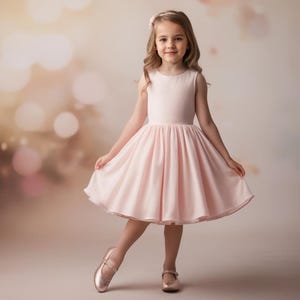 以下が含まれることがあります： ピンクの袖なしのドレスを着た若い女の子が、スカートをいっぱいに広げて立っています。彼女はぼかし効果のある薄い茶色の背景に立っています。