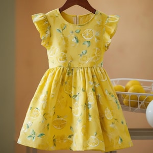 May include: A yellow dress with a lemon print. The dress has short, ruffled sleeves and a fitted bodice that flows into a flared skirt. The lemon pattern includes slices and leaves. The dress is hanging on a wooden hanger.