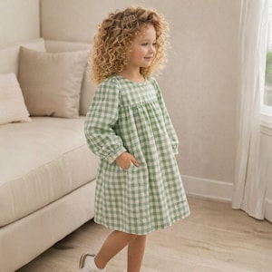 May include: A young girl wearing a green and white gingham dress with long sleeves and pockets. The dress has a round neckline and a loose, comfortable fit. She is wearing white shoes and has curly blonde hair.
