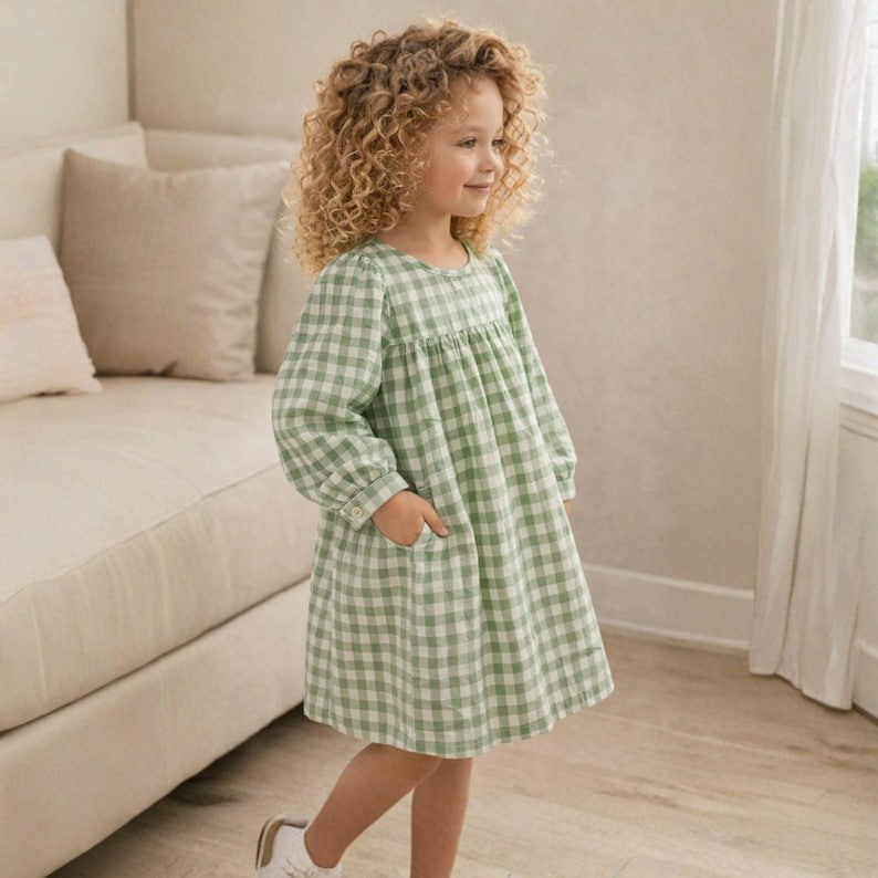 May include: A young girl wearing a green and white gingham dress with long sleeves and pockets. The dress has a round neckline and a loose, comfortable fit. She is wearing white shoes and has curly blonde hair.