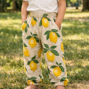 May include: Beige trousers featuring a pattern of yellow lemons and green leaves. The trousers have an elasticated waistband and pockets. The fabric looks like a lightweight linen or cotton blend. The trousers are designed for children.