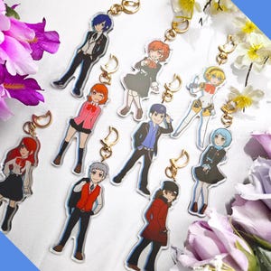 May include: A selection of anime-style character keychains. Each keychain showcases a unique character design with bright colours and detailed artwork. The keychains are attached to gold-coloured metal clasps.