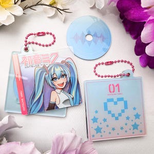 May include: A collection of Hatsune Miku themed keychains and a circular coaster. The keychains feature anime artwork and the text "Hatsune Miku". One keychain has a pixelated heart design and stars. The coaster has a sound wave design.