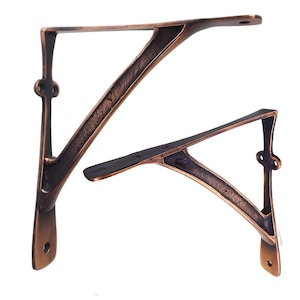 Könnte beinhalten: Zwei bronzefarbene Regalhalterungen. Die Halterungen haben ein dekoratives Design mit einem gebogenen Stützarm und einer flachen Oberseite. Jede Halterung hat vorgebohrte Löcher zur Montage. Die Oberfläche hat ein rustikales, antikes Aussehen.