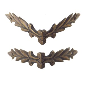 May include: Two antique bronze-colored corner brackets with a leaf and fleur-de-lis design. The decorative metal hardware is in a symmetrical, angled shape, perfect for furniture or craft projects.