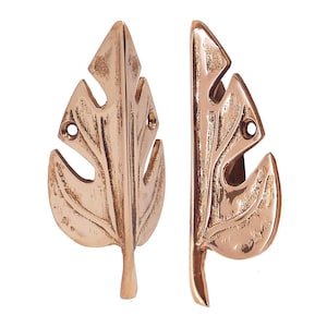 May include: Two bronze-coloured leaf-shaped cabinet pulls. Each pull has a detailed leaf vein design and two holes for mounting. The pulls are made of metal and have a rustic, antique appearance. They are designed for use on cabinets or drawers.