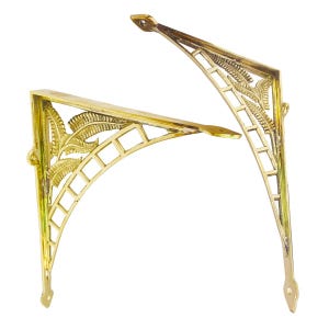 Puede incluir: Dos soportes de estante de metal dorado con un diseño decorativo de hojas y un patrón de cuadrícula. Los soportes están en ángulo y tienen una punta puntiaguda en el extremo.