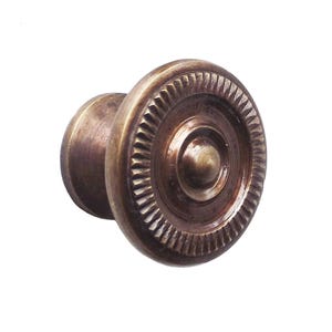 May include: A bronze-colored cabinet knob with a circular design. The knob features a central, raised button surrounded by concentric rings and a textured outer edge. The hardware is likely for furniture or cabinetry.