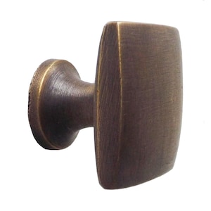 Może przedstawiać: Mosiężny uchwyt do szafki z prostokątną powierzchnią i zaokrągloną podstawą. Uchwyt ma szczotkowaną metalową fakturę. Ten osprzęt nadaje się do szuflad i szafek.