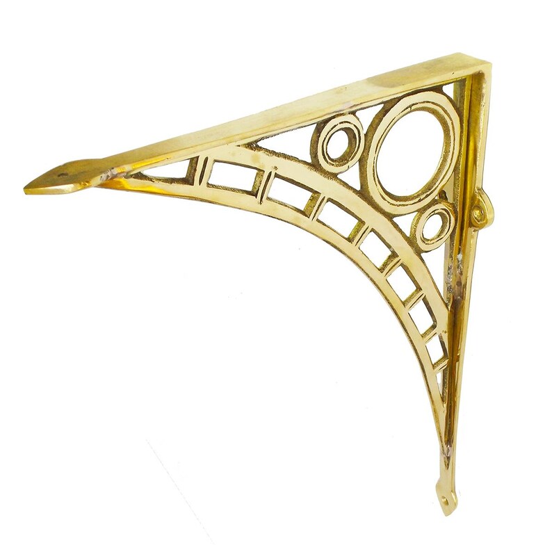 May include: A gold-colored, Art Deco-style shelf bracket. The bracket has a triangular shape with a decorative openwork design featuring circles and rectangular cutouts. The bracket is made of metal and has a small lip at the end.