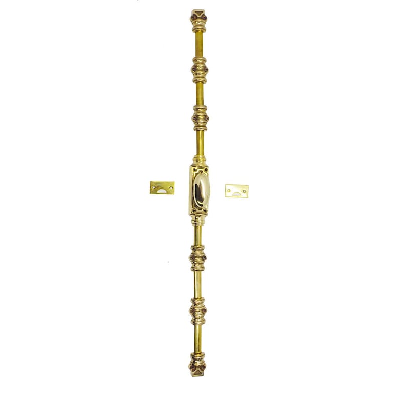 May include: A vertical brass door bolt with decorative detailing. The bolt features an oval center piece, multiple ornate rings, and square end caps. Two small brass plates are included. The hardware is designed for door security and adds a touch of elegance.