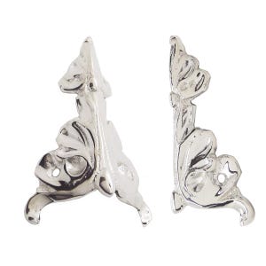 May include: Two silver-colored decorative furniture corner brackets. Each bracket has a detailed, ornate design with a curved base, a central body with a leaf-like pattern, and a top section with a similar design. The brackets are made of metal.
