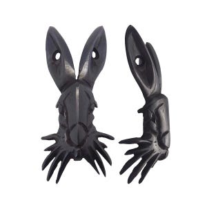 Puede incluir: Dos esculturas de cabeza de conejo de madera tallada de color gris oscuro. Las cabezas de conejo tienen orejas largas con un agujero en cada oreja. Las cabezas de conejo tienen una cara detallada y un diseño puntiagudo en la parte inferior.