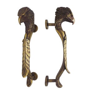 Może przedstawiać: Dwa mosiężne uchwyty do drzwi, każdy wyrzeźbiony w kształcie głowy orła. Uchwyty mają szczegółowe wzory piór i dziobów. Uchwyty są zamontowane na białym tle, prezentując ich skomplikowane rzemiosło.