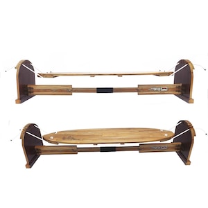 May include: Two wooden surfboard racks. Each rack has a dark brown curved end piece with white rope. The surfboard rests on a light brown wooden base. The base is connected to a wooden frame with a black strap.