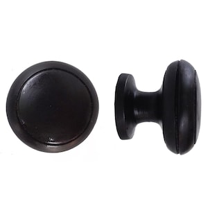 Könnte beinhalten: Zwei dunkelbraune Schrankknöpfe. Einer ist eine runde, flache Scheibe mit leicht erhöhtem Rand. Der andere ist ein zylindrischer Knopf mit einem abgerundeten, geriffelten Griff. Beide bestehen aus einem festen, dunklen Material.