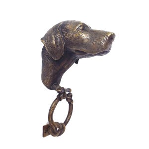 Op de afbeelding: Een bronzen deurklopper in de vorm van een hondenkop met een ring aan de onderkant. De hondenkop is naar rechts gericht en de ring is in de vorm van een knoop.