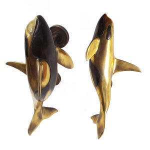 May include: A pair of brass door handles shaped like orcas, with black accents. The handles are shown from the side, with the tail fin facing the viewer.