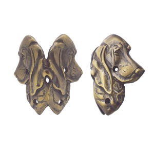 May include: Two antique brass-toned decorative dog head corner brackets. One bracket features two dog heads, and the other has a single dog head. Each has pre-drilled holes for mounting. The dog heads have detailed features.